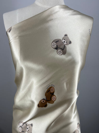 A pale Printed Satin - Teddy - 150cm fabric from Super Cheap Fabrics draped over a mannequin is adorned with small prints of teddy bears. The teddy bears are in various shades of brown, each holding a small heart that reads "Teddy Bear." This lightweight fabric has a slight sheen, adding a touch of elegance.