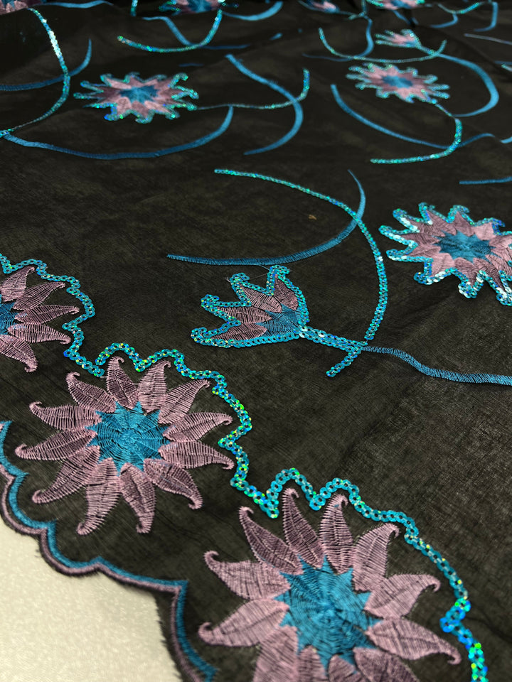 A close-up of Super Cheap Fabrics' Embroidered Sequins - Blue Stars - 130cm adorned with intricate floral embroidery. The 100% polyester design features pink and blue flowers accented with shiny, teal sequins, creating a vibrant and eye-catching pattern. This 75 GSM fabric's edge is scalloped with the same multi-colour floral motif.