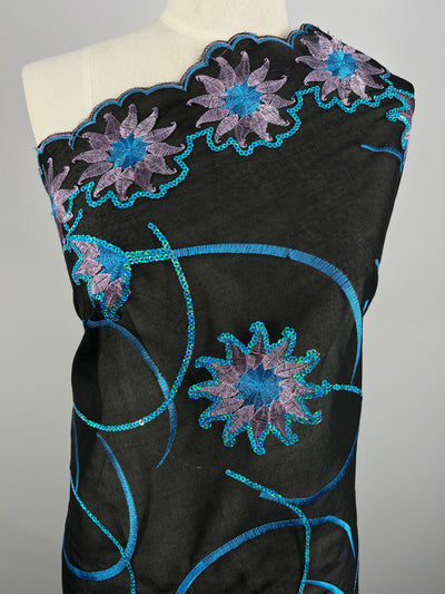 A mannequin draped in black Embroidered Sequins - Blue Stars - 130cm fabric from Super Cheap Fabrics embroidered with intricate, multi-colour blue and purple floral designs and swirling blue patterns. The flowers have petals outlined in blue and filled with purple, creating a vibrant and elegant look against the dark background.