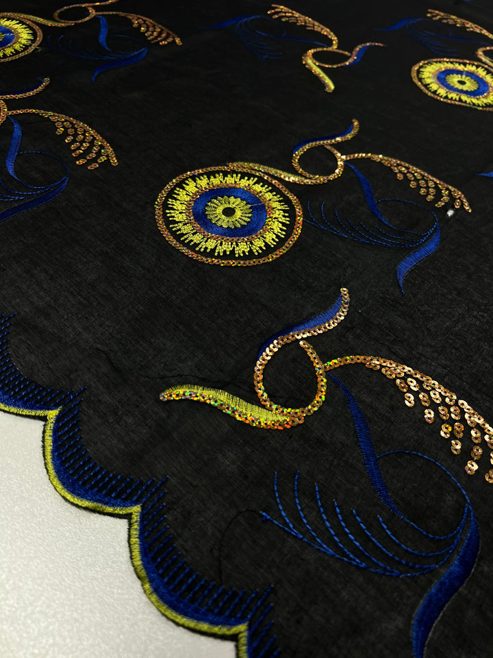 A piece of black fabric, 100% polyester and 75 GSM, is adorned with intricate gold and blue embroidery. The design features multi-colored circular floral patterns with surrounding curvy, leaf-like elements and reflective sequins, creating an elegant and detailed appearance. The fabric has a scalloped edge. This is the Embroidered Sequins - Bright Eye - 130cm from Super Cheap Fabrics.