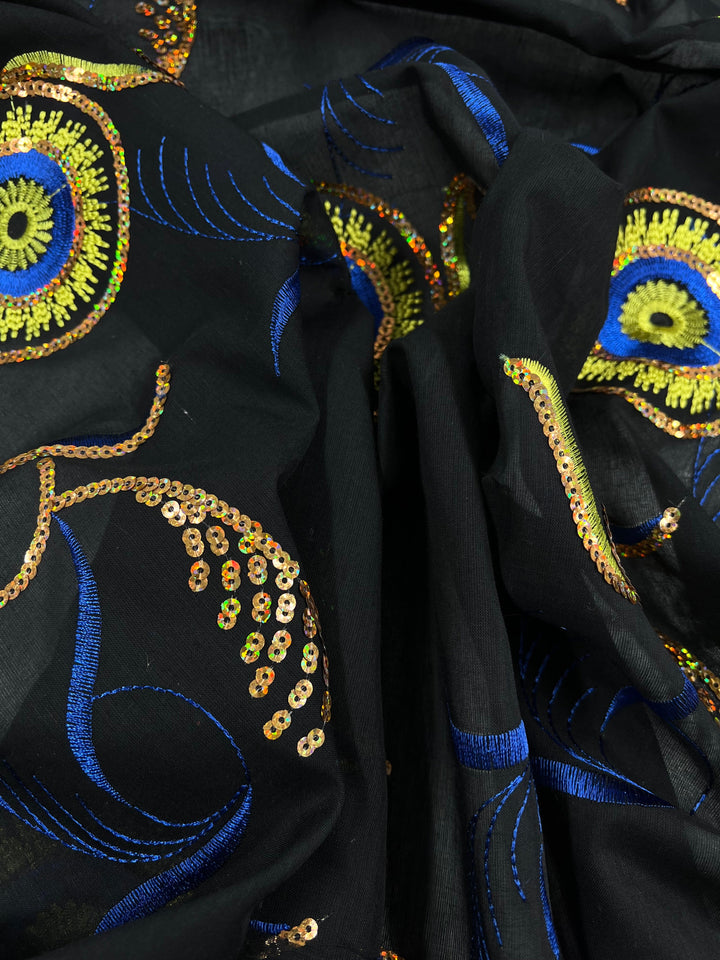 Close-up of a black, 100% polyester fabric featuring intricate, colorful embroidery with gold sequins. The design includes blue swirls and bright yellow circles, giving a peacock feather-like appearance. The **Super Cheap Fabrics Embroidered Sequins - Bright Eye - 130cm** is gathered, showcasing its texture and detailed embroidery work.