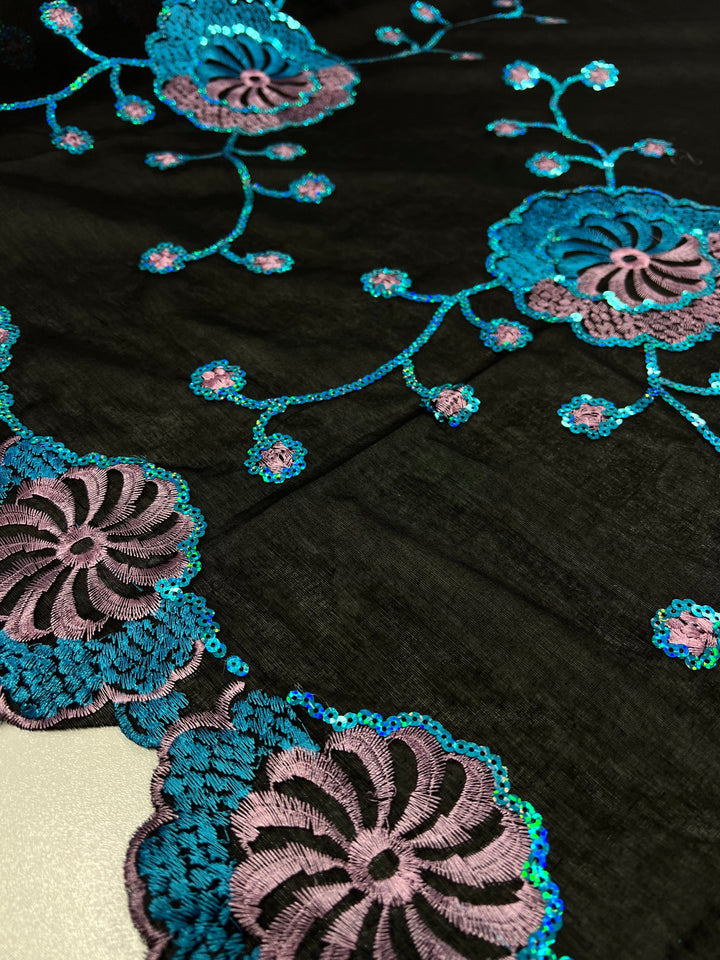 Close-up of a black, 100% polyester fabric adorned with intricate teal and pink floral embroidery. The multi-colour flowers and trailing stems are detailed with sequins, creating a shimmering effect. The delicate GSM 75 fabric is richly textured, showcasing the elaborate needlework. This is the Embroidered Sequins - Blurple Clusters - 130cm by Super Cheap Fabrics.
