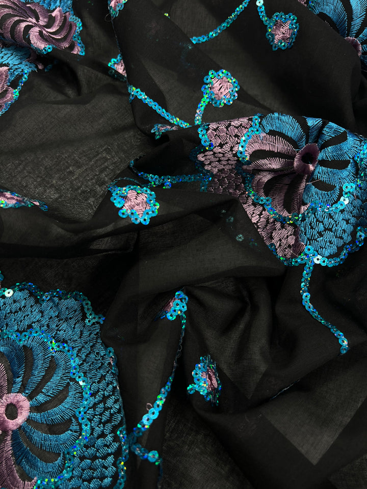 A close-up view of a piece of black fabric adorned with intricate embroidery. The Super Cheap Fabrics Embroidered Sequins - Blurple Clusters - 130cm features teal and purple floral patterns with sequined accents, creating a vibrant, textured design on the dark background. Crafted from 100% polyester for durability and sheen.