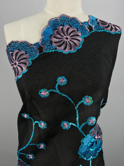 A black fabric draped over a mannequin features intricate, multi-color floral embroidery. The Super Cheap Fabrics Embroidered Sequins - Blurple Clusters - 130cm is done in blue and purple, forming large flowers at the top and bottom edges with smaller blossoms on winding stems across the 100% polyester fabric.