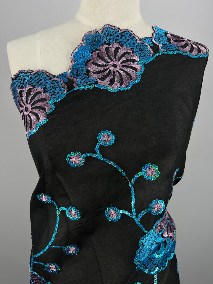 A mannequin is draped in Super Cheap Fabrics' Embroidered Sequins - Blurple Clusters - 130cm, adorned with intricate blue and purple floral embroidery. The design features large flowers along the top edge and smaller blossoms connected by winding stems throughout the GSM 75 material, creating an elegant, ornamental pattern.