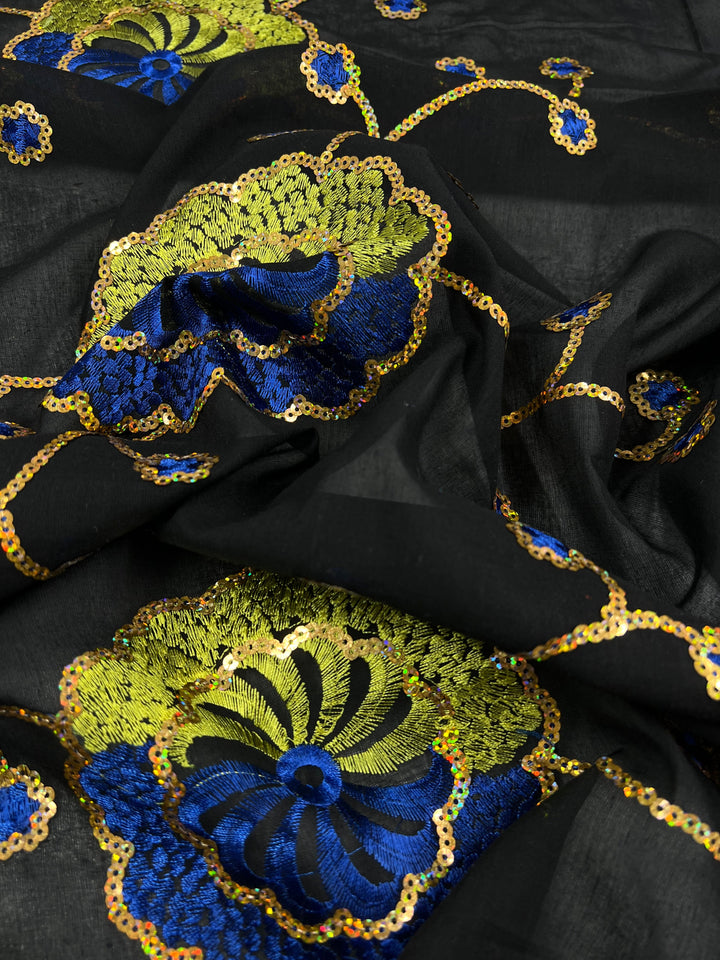 Close-up of Super Cheap Fabrics Embroidered Sequins - Clusters - 130cm width, multi-colour fabric embellished with intricate, colorful embroidery. The design features large flower-like patterns in vibrant blue and green, highlighted with shimmering golden sequins that catch the light, creating a striking contrast against the dark polyester background.