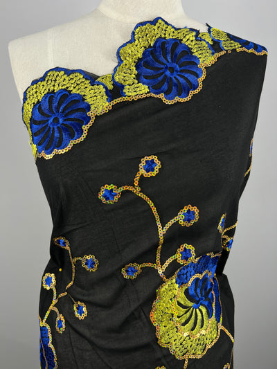 A close-up of a mannequin draped with Embroidered Sequins - Clusters - 130cm by Super Cheap Fabrics. The detailed polyester embroidery features large blue flowers with yellow accents and gold outlining, creating a vibrant design against the dark background.