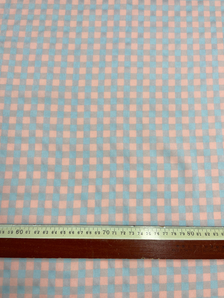 A medium weight fabric with a checkered pattern in light blue and pink squares. Made from a cotton polyester blend, this textile is perfect for clothing use. A wooden ruler with a metal edge is placed horizontally across the bottom of the image, indicating the scale of the pattern. Introducing Super Cheap Fabrics' Cotton Poly - Pale Pink & Blue Gingham - 150cm.