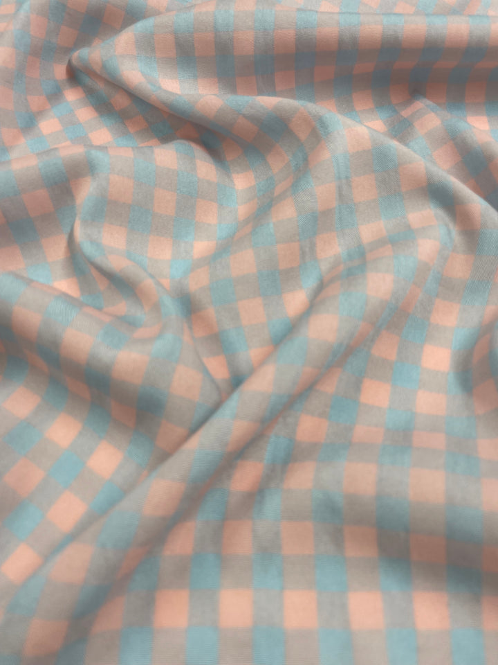 Close-up image of a medium weight fabric with a checkered pattern, featuring alternating light pink and blue squares. Perfect for clothing use, the Cotton Poly - Pale Pink & Blue Gingham - 150cm by Super Cheap Fabrics lays in soft folds with a slightly textured appearance, creating a sense of depth and gentle movement.