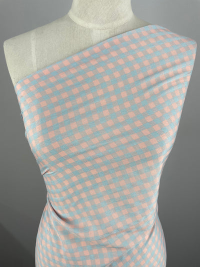 A mannequin is draped with Super Cheap Fabrics' Cotton Poly - Pale Pink & Blue Gingham - 150cm. The cotton polyester blend is arranged in a one-shoulder style, emphasizing the gentle folds and texture, perfect for clothing use.