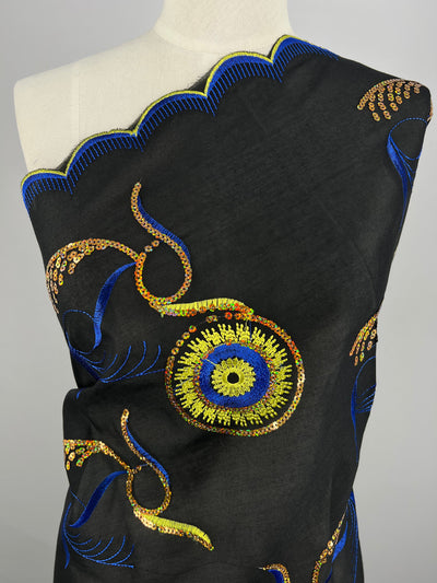 A mannequin is draped in Super Cheap Fabrics' Embroidered Sequins - Bright Eye - 130cm, featuring intricate embroidery. The design includes blue, yellow, and orange sequined circular patterns and swirling lines. The top edge of the fabric has a scalloped blue border.