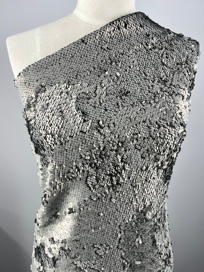 A close-up of a mannequin wearing an asymmetrical, one-shoulder garment covered in Super Cheap Fabrics' Premium Sequins - Matte Silver - 150cm. The sequins create a shimmering, textured surface that catches the light, giving the heavy weight fabric a sparkling, metallic appearance. The background is plain white.