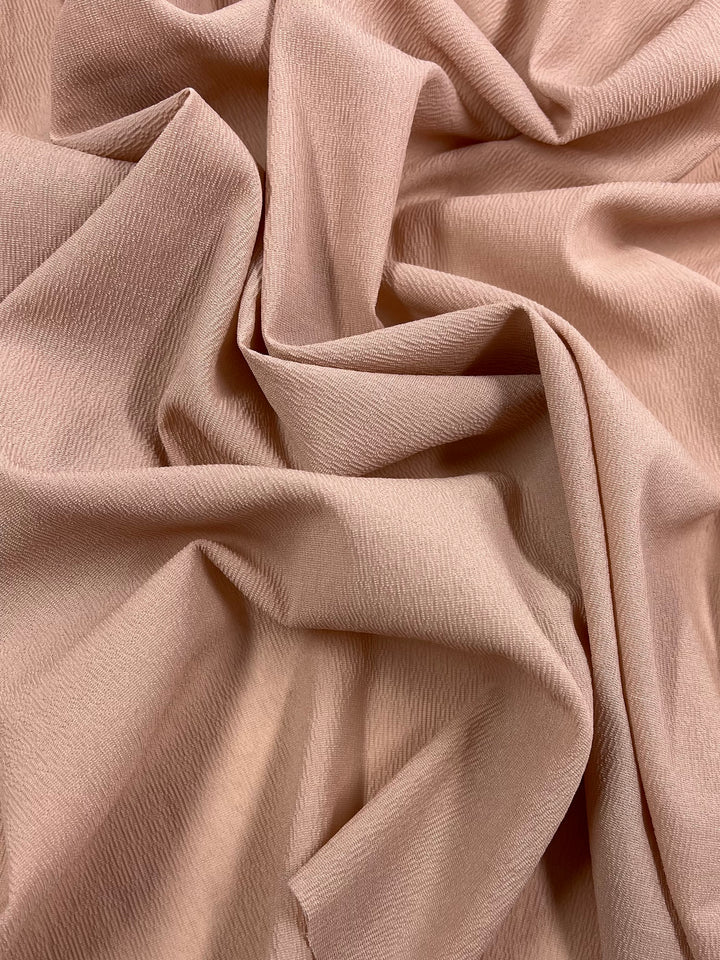 A close-up shot showcases a fabric with a soft, smooth texture. The material, named Polyester Broad Cloth - Misty Rose from the brand Super Cheap Fabrics, exhibits a gentle sheen. It is loosely gathered in folds and drapes, creating natural shadows and highlights.