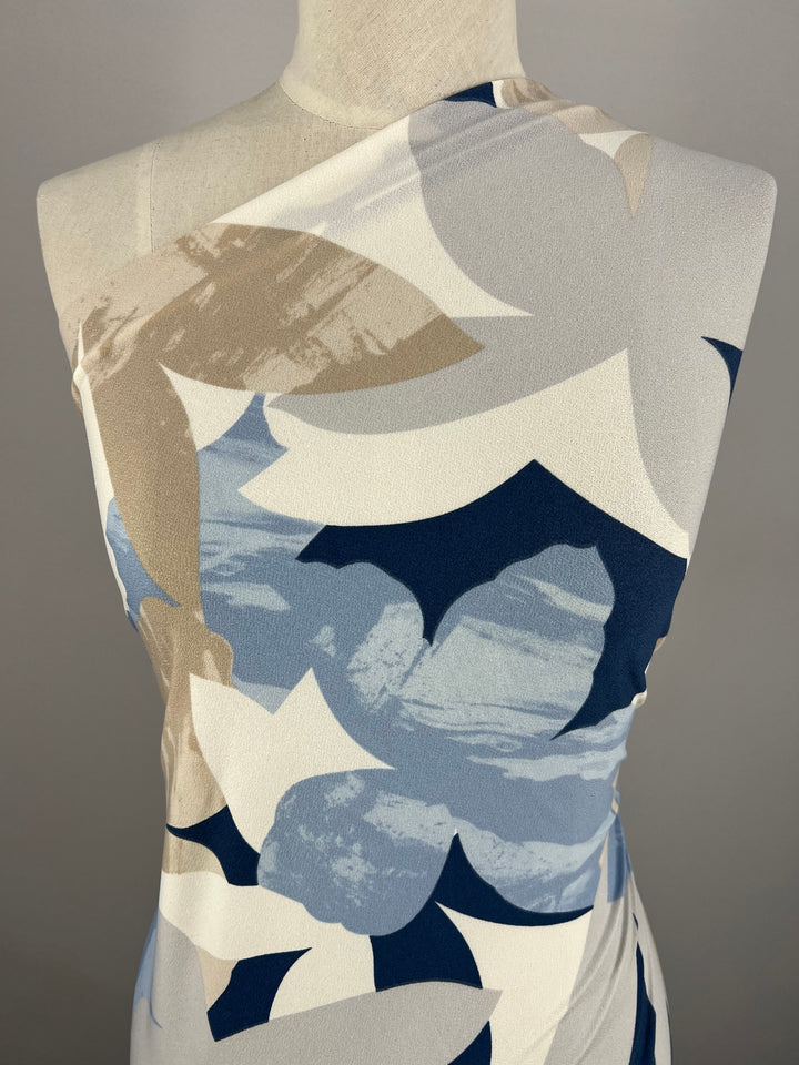 A mannequin dressed in a sleeveless, asymmetrical top crafted from medium-weight Polyester/Spandex fabric with a large floral pattern in shades of blue, white, and beige stands against a neutral gray background using Super Cheap Fabrics' Printed Lycra - Flower Projection - 150cm.