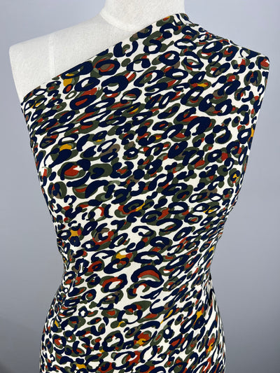 A mannequin wearing a form-fitting one-shoulder dress crafted from Super Cheap Fabrics' Printed Lycra - Painters Palette - 150cm with a colorful leopard print pattern, featuring shades of navy blue, red, olive green, and beige on a white background. The dress includes two-way stretch and the mannequin is set against a plain gray backdrop.