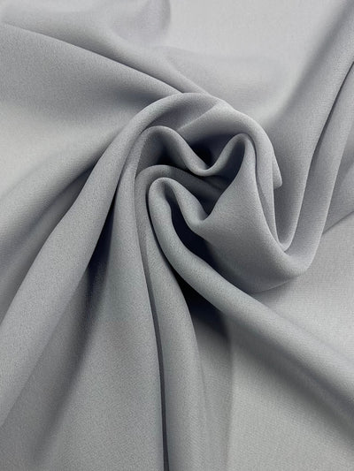 A close-up of Super Cheap Fabrics' Plain Georgette - Grey Dawn - 112CM, a light grey, 100% polyester fabric that is softly draped and gently folded in the center, creating smooth curves and subtle shadows across its textured surface.