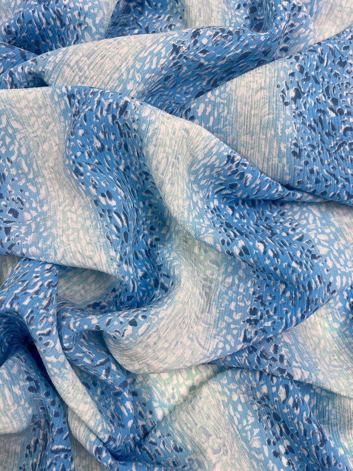 A close-up of light, airy polyester fabric with a soft, abstract pattern. The material features various shades of blue and white, creating a textured effect that resembles brush strokes or scattered spots. This multi-use fabric appears to be lightweight and flowing. The product shown is Designer - Droplet Stripes - 150cm by Super Cheap Fabrics.