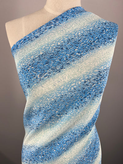 A mannequin is draped in the Designer - Droplet Stripes - 150cm polyester fabric by Super Cheap Fabrics, featuring a light blue and white striped pattern with a textured, lacy design. The lightweight fabric wraps over one shoulder, showcasing its airy and delicate look. The neutral background highlights the intricate details of this multi-use material.