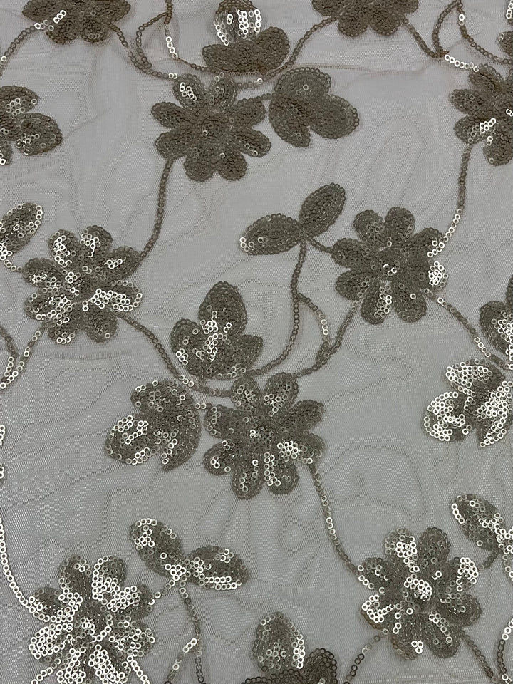 A close-up view of a fabric with an embroidered floral pattern. The flowers are adorned with metallic sequins, adding a shimmering effect to the Designer Sequins- Gold Calendula - 150cm by Super Cheap Fabrics. The background material is transparent and lightweight, giving a delicate and elegant appearance.