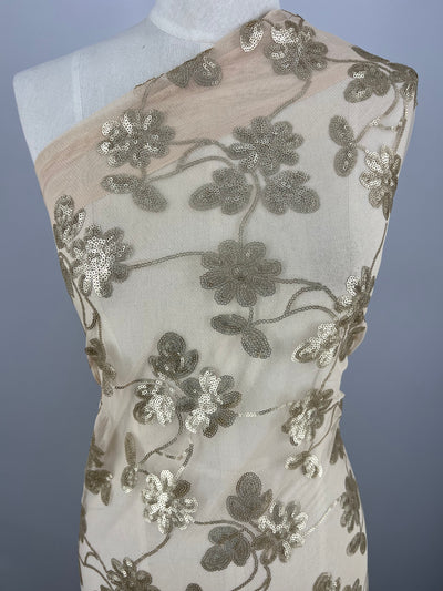 A white mannequin is draped with a sheer beige Designer Sequins- Gold Calendula - 150cm fabric from Super Cheap Fabrics, adorned with intricate gold floral embroidery. The arrangement of flowers and leaves is detailed, sparkling under the light, creating an elegant and luxurious appearance. The background is plain gray.