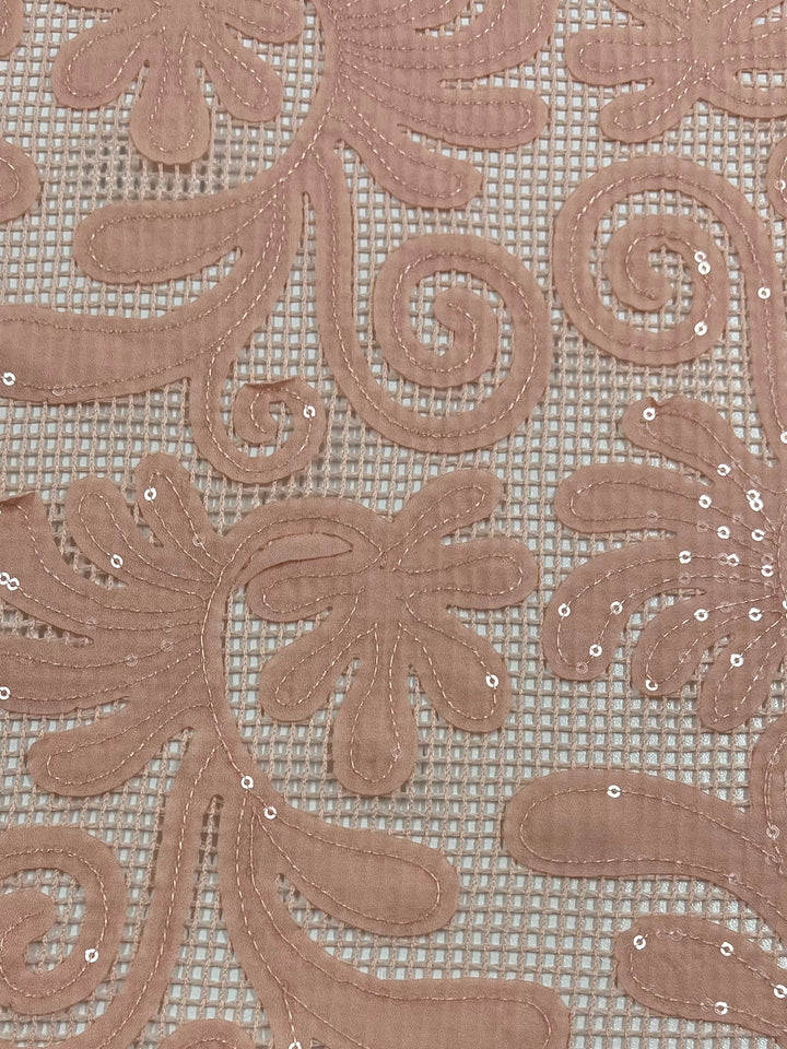 A close-up image of Super Cheap Fabrics' Embellished Netting - Peach Splash - 150cm, with a delicate pattern of swirling lines, floral shapes, and sequins. The lace is beige and set against a light, transparent mesh background, creating an elegant and textured appearance ideal for formal wear.