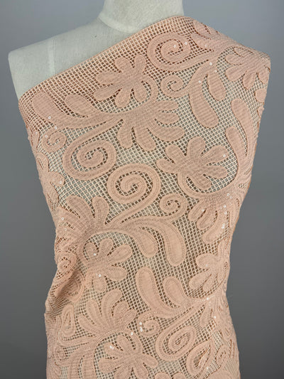 A beige one-shoulder dress on a mannequin, featuring an intricate, swirling floral lace pattern with sequins embellishments made from Embellished Netting - Peach Splash - 150cm by Super Cheap Fabrics. The lace overlay, made from polyester fabric, highlights the delicate and intricate design, creating a stylish and elegant formal wear piece.