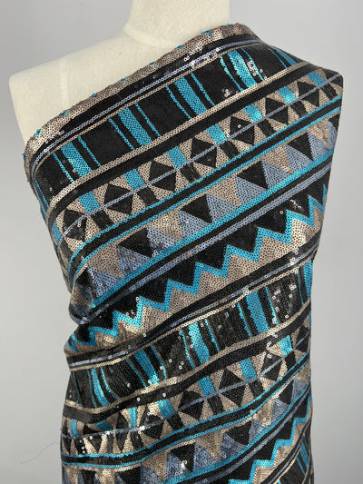 A lightweight fabric draped over a mannequin, featuring a geometric pattern with black, teal, silver, and beige colors. The Designer Sequins- Stripe Zag - 150cm by Super Cheap Fabrics is adorned with sequins, creating a shimmering effect. The design includes zigzag, triangle, and linear motifs—ideal for dance wear.