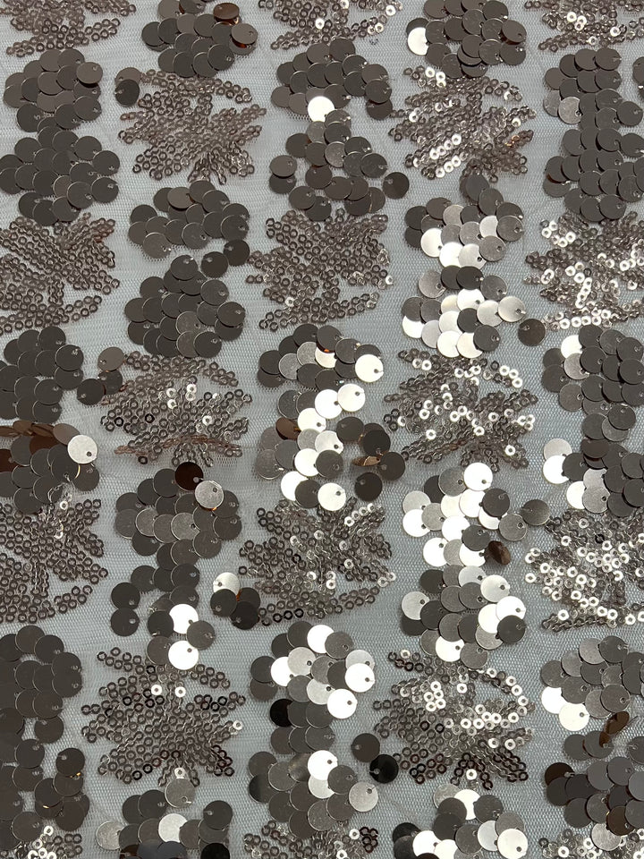 A close-up view of a lightweight fabric adorned with silver sequins arranged in intricate, fern-like patterns. The shiny sequins create a textured, reflective surface that catches the light, set against a light-colored background. Product Name: Designer Sequins- Rose Gold Canopy - 150cm Brand Name: Super Cheap Fabrics