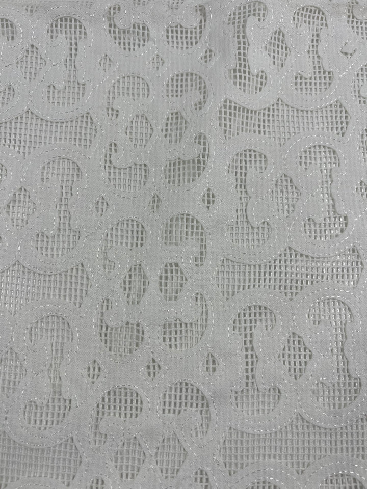 Close-up of a white lace fabric featuring intricate, swirling patterns. The design includes a combination of solid and mesh sections, creating a delicate, decorative texture. This lightweight fabric appears to be airy and is 100% polyester, perfect for designer embellished netting projects. The product being described is the Embellished Netting - Heritage - 150cm from Super Cheap Fabrics.