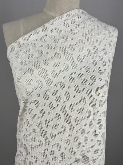 A torso draped in Embellished Netting - Heritage - 150cm by Super Cheap Fabrics features delicate, semi-transparent white lace fabric with an intricate floral pattern. The 100% polyester lace is soft with an elegant design, displayed on a light beige mannequin against a neutral gray background.