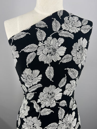 A dress on a mannequin featuring a black background with a white floral pattern. The dress, made from Super Cheap Fabrics' Printed Lycra - White Camellia - 150cm fabric, has an asymmetrical, one-shoulder design with detailed white flowers and leaves throughout. The plain grey background highlights its intricate design and two-way stretch comfort.