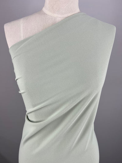 A dress form displays a light green, one-shoulder dress made of Plain Scuba Crepe - Sea Foam - 160cm by Super Cheap Fabrics. The fabric drapes gracefully from the shoulder across the bust and gathers slightly at the side, creating soft folds. The medium weight fabric against a plain gray background highlights the dress's simple elegance.