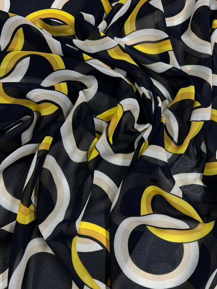 A close-up of a fabric with a bold, geometric pattern. The design features intertwined rings in yellow, white, gray, and black on a dark background, creating a dynamic and layered visual effect. Ideal for sewing projects, this lightweight Printed Chiffon - Links - 160cm material from Super Cheap Fabrics appears slightly crumpled.