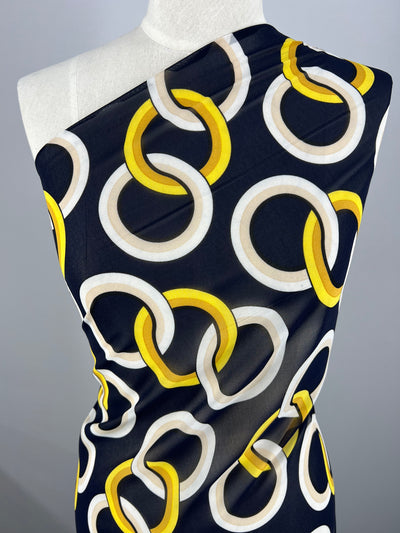 A mannequin draped with Super Cheap Fabrics' Printed Chiffon - Links - 160cm featuring a bold pattern of interlocking rings in white, grey, and yellow. The polyester chiffon fabric covers the upper torso and has a smooth texture, showcasing the modern and vibrant design against a plain, grey background—perfect for sewing projects.