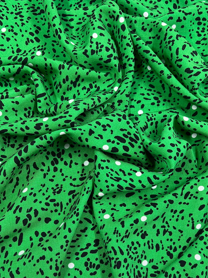 A close-up image of a green, lightweight fabric with a pattern of small black and white spots. The Super Cheap Fabrics Bamboo Rayon - Dotted Safari - 150cm is draped in loose folds, showcasing its texture and design, making it ideal for stylish home decor.