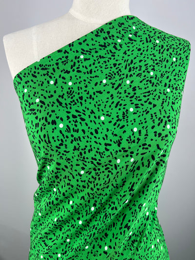 A one-shoulder garment on a mannequin, featuring a bright green lightweight fabric with a pattern of black and white speckles. The fabric is draped smoothly over the mannequin, highlighting the texture and color of the material. The background is a neutral gray, perfect for any home decor setting. This beautiful fabric is Bamboo Rayon - Dotted Safari - 150cm by Super Cheap Fabrics.
