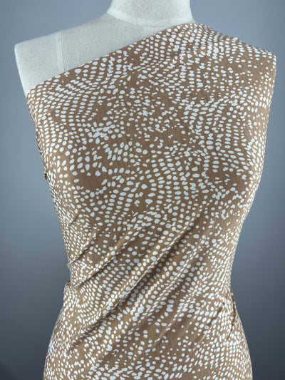 A dress mannequin is draped in a one-shoulder brown medium-weight fabric, featuring an intricate white dotted pattern resembling scattered seeds or pebbles. The background is a plain gradient from light to dark gray. The fabric used is Printed Lycra - Sirocco Reptilian - 150cm by Super Cheap Fabrics.