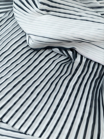 A close-up shot of lightweight fabric with black and ivory vertical stripes. The 100% polyester material is slightly crumpled, creating wavy, uneven lines. The texture and folds of the fabric add depth and dimension to the image. This is the Pleated - Black & Ivory - 145cm from Super Cheap Fabrics.