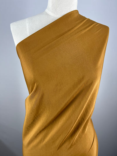 A dress form is wrapped in a smooth, Microfibre - Brown Sugar - 150cm fabric from Super Cheap Fabrics, draped elegantly over one shoulder in a diagonal fashion. The material appears soft and slightly lustrous, creating gentle folds and contours. The background is plain gray.