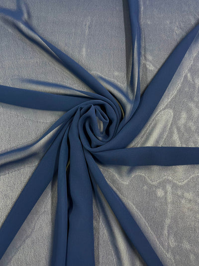 A piece of Hi-Multi Chiffon - Navy - 150cm by Super Cheap Fabrics is artistically arranged in a spiral pattern on a gray surface, displaying its smooth texture and flowing drape. The sheer material appears slightly translucent, with a soft sheen under the light.