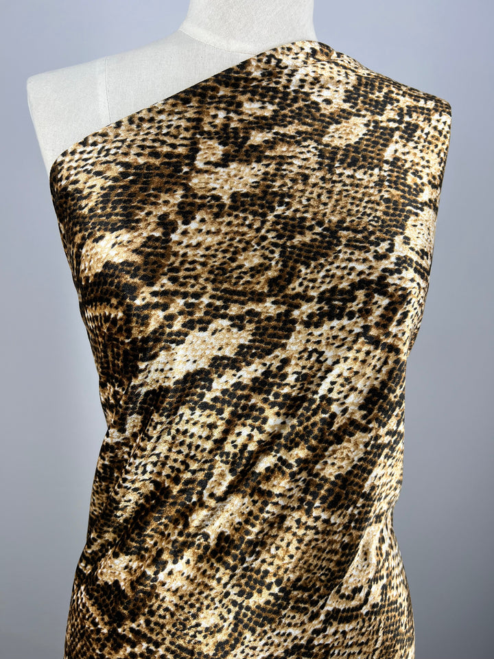 A white mannequin displays a one-shoulder dress in Super Cheap Fabrics’ Printed Velvet - Python, a light to medium weight polyester with a brown, black, and beige snakeskin pattern. The fabric is 150cm wide. Background is plain and neutral.