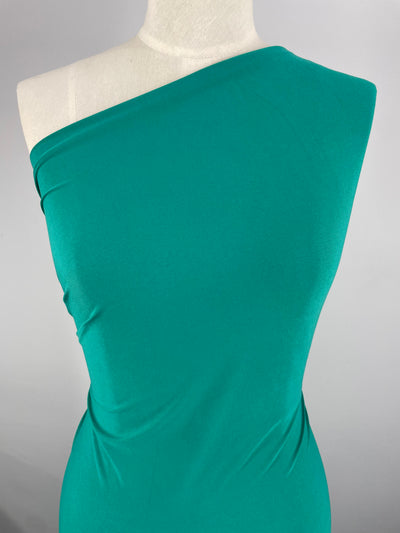 A close-up image of a dress form wearing a vibrant teal, one-shoulder dress made from medium weight fabric. The smooth and fitted material drapes elegantly over the form, showcasing ITY Knit - Sea Green - 150cm by Super Cheap Fabrics. The background is plain and minimal, emphasizing the dress's color and design.