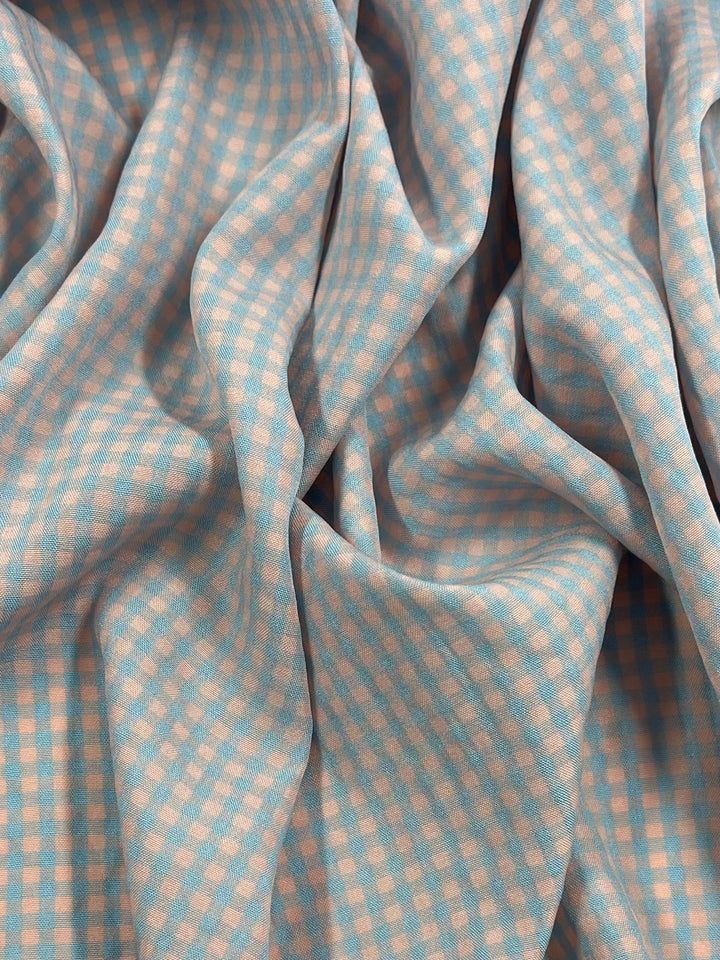 A close-up of a light weight fabric with a blue and light peach checkerboard pattern, showcasing its soft, draped texture with folds and creases. Made from a cotton polyester blend, the small squares create a subtle, uniform design. Introducing Cotton Poly - Mini Gingham Pale Pink & Blue - 150cm by Super Cheap Fabrics.