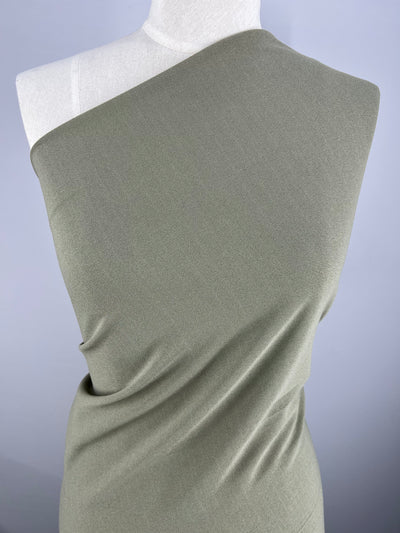 A close-up view showcases a dress form draped in Super Cheap Fabrics' Plain Scuba Crepe - Laurel Oak - 170cm fabric. This stretchy fabric boasts a smooth texture, styled in an asymmetrical one-shoulder design. The plain and light-colored background highlights both the fabric and the garment design beautifully.