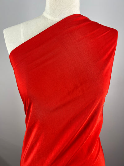 A mannequin draped in **Super Cheap Fabrics' Microfibre - Red - 150cm**, wrapped over one shoulder, creates an elegant, asymmetrical look. The background is plain and gray, highlighting the vivid color and smooth texture of the 100% polyester material.