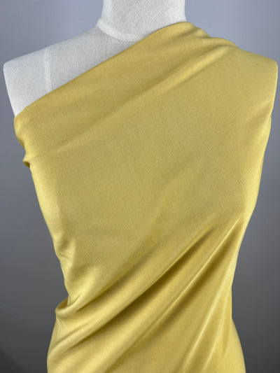 A mannequin is draped with a piece of smooth, mustard-yellow Microfibre - Rattan - 150cm from Super Cheap Fabrics that is wrapped around its torso in an asymmetrical style, covering one shoulder and under the opposite arm. The background is plain and gray, highlighting the lightweight fabric's color and texture.
