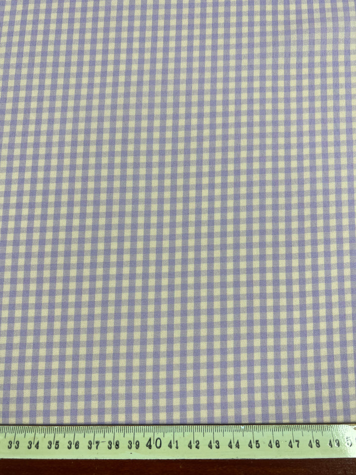 A piece of light weight fabric with a small purple and white checkered pattern is shown. At the bottom of the image, a green measuring tape is placed horizontally, indicating the width of this versatile, multi-use Cotton Poly - Mini Gingham Peach & Purple - 150cm fabric from Super Cheap Fabrics.