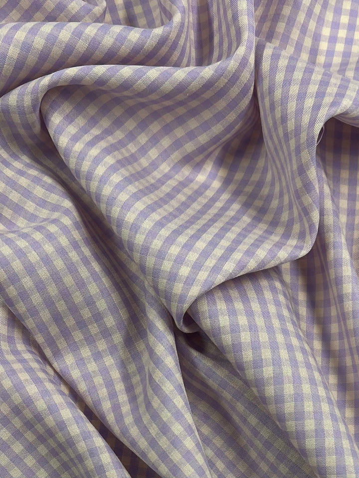 A close-up shot of crumpled light purple and white gingham fabric with a soft and smooth texture. The small checkered pattern creates a subtle contrast, and the Cotton Poly - Mini Gingham Peach & Purple - 150cm from Super Cheap Fabrics catches light at various angles, creating gentle shadows and highlights.