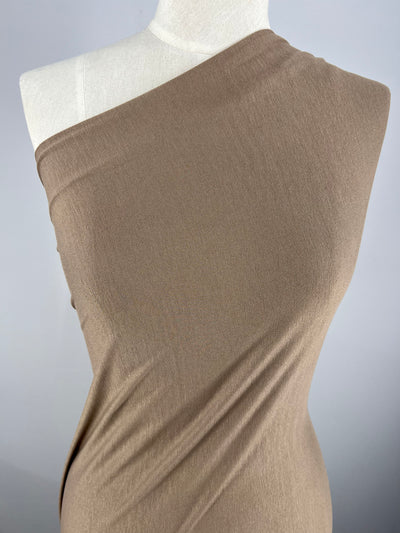 A close-up image of a dress form wrapped in a piece of light brown, one-shoulder medium weight fabric. The Rayon Lycra - Coca Mocha - 150cm material from Super Cheap Fabrics appears soft and drapes smoothly over the mannequin, creating a simple and elegant appearance against a plain gray backdrop.