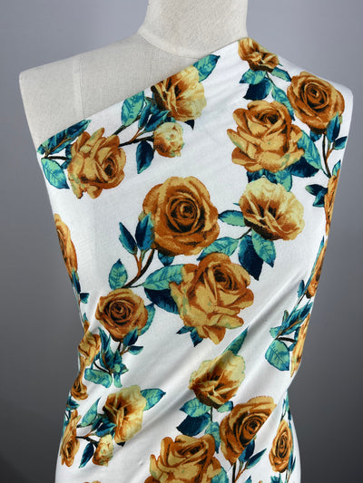 A mannequin draped with **Super Cheap Fabrics' Printed Rayon - La Flora - 145cm**. The lightweight fabric covers one shoulder and wraps diagonally, showcasing the yellow-orange roses and green leaves floral pattern against a gray background.